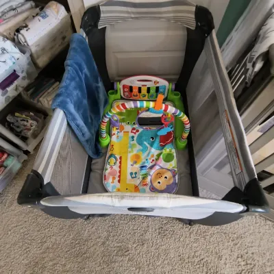 Graco Pack 'n Play portable playard containing a Fisher-Price Deluxe Kick & Play Piano Gym and a blue plush blanket.