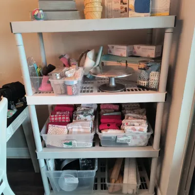 Four-tier white plastic shelving unit filled with baking supplies, including a Cuisinart appliance, cake pans, a rotating cake stand, and...