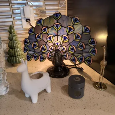 Tiffany-style stained glass peacock accent lamp, shown illuminated alongside a white ceramic llama planter, a green ceramic tree, and a...