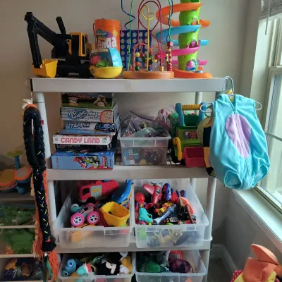 A white plastic shelving unit filled with various children's toys, board games including Candy Land, plush animals in clear bins,...