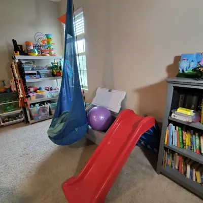 Children's playroom items including a red plastic slide, a hanging blue sensory swing, a purple exercise ball, and a grey bookshelf...