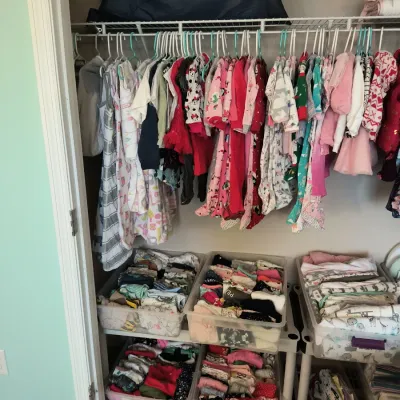 Assorted baby and toddler clothing hanging in a closet and organized in clear plastic storage bins below, featuring various colors and...