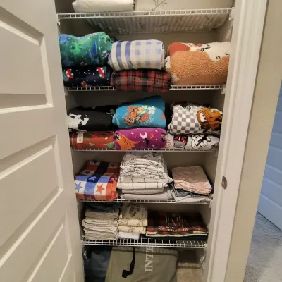 Assorted household linens, blankets, and sheets, including themed children's blankets, neatly folded on wire closet shelving.