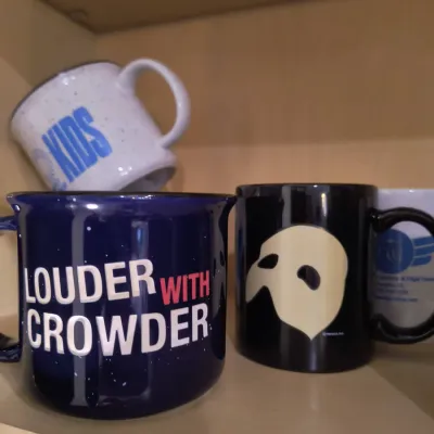 Collection of novelty coffee mugs, including a blue "Louder with Crowder" campfire-style mug and a black Phantom of the Opera...
