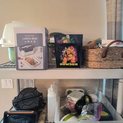Assorted household items on shelving, including a boxed electric lunch box, Super Mario themed tin containers, a woven basket, a cast...