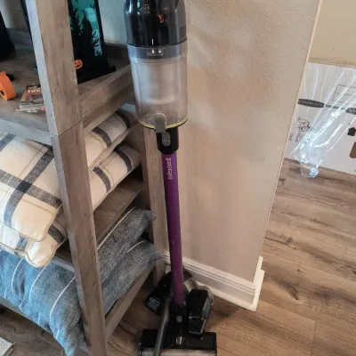 Bissell cordless stick vacuum cleaner with a purple wand and black motor housing, shown standing next to a wooden shelving unit.