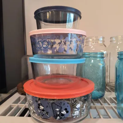 Stack of Pyrex glass food storage containers with plastic lids, including Disney-themed designs, displayed alongside vintage blue and...