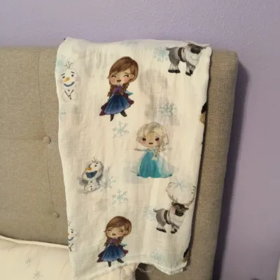 Disney Frozen themed muslin swaddle blanket draped over a beige tufted headboard, featuring chibi-style illustrations of Anna, Elsa,...