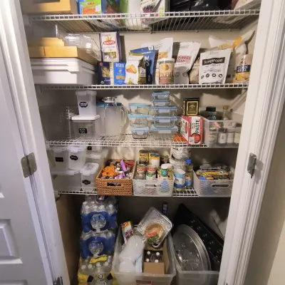 Pantry closet stocked with various dry goods, canned foods, storage containers, bottled water, and kitchen organization bins.