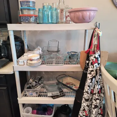 White plastic four-tier shelving unit holding various kitchenware, including Pyrex storage containers, blue Mason jars, a pink mixing...