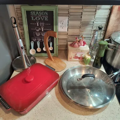 Assorted kitchenware including a red enameled cast iron baker, a stainless steel skillet with glass lid, a wooden banana hanger, and...