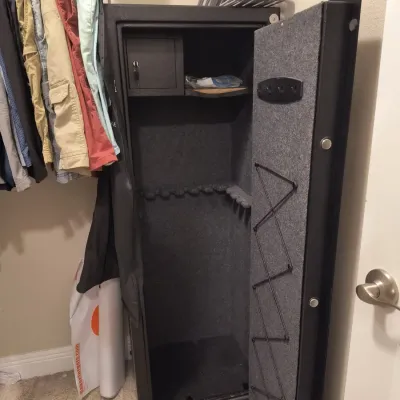 Tall black metal gun safe with a carpeted interior, featuring an upper locking compartment and door storage, shown open in a closet.