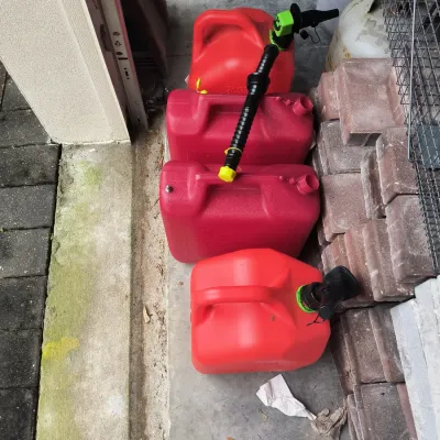 Four red plastic gas cans of varying sizes, some with spouts attached, stored outdoors next to a stack of bricks.
