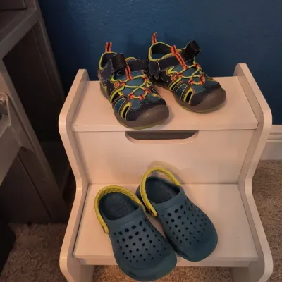 Two pairs of children's shoes, including blue and green clogs and colorful athletic sandals, displayed on a white wooden step stool.