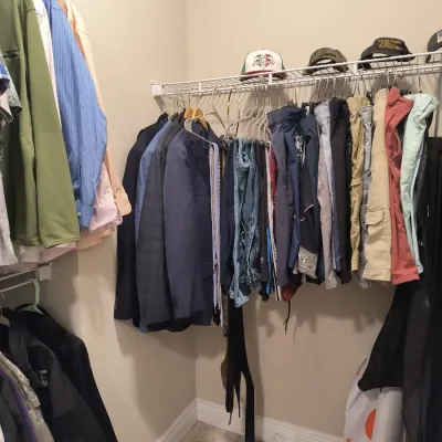 Assorted men's clothing including suit jackets, jeans, trousers, and button-down shirts hanging in a closet, accompanied by baseball...