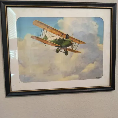 Framed vintage illustration of a green and yellow biplane flying through clouds, signed by the artist in the lower right corner.