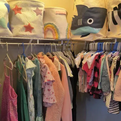 Assorted children's clothing hanging on a wire closet rack, topped with decorative fabric storage bins featuring stars, rainbows,...
