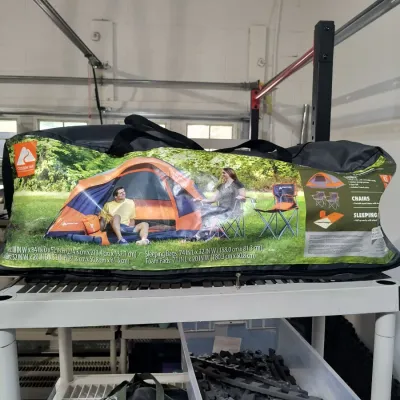 Ozark Trail camping combo set in its original carrying bag, including a tent, chairs, and sleeping pads, resting on a white wire shelf.