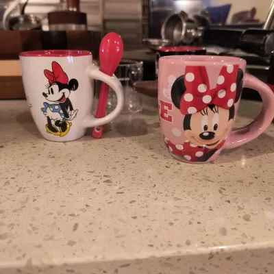 Two Disney Minnie Mouse ceramic mugs, including a white mug with a matching red spoon and a pink mug featuring a close-up of Minnie.