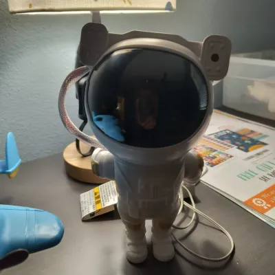 Modern astronaut-shaped galaxy star projector lamp with a white plastic body and dark visor, shown on a desk next to a table lamp.
