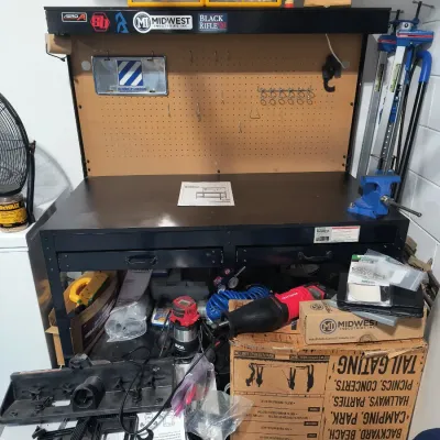 Black metal workbench with integrated pegboard back, overhead shelf, and dual drawers, shown with various tools and accessories including...