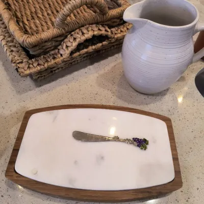 Marble and wood cheese board with a grape-themed spreader, shown alongside a white ceramic pitcher and woven baskets.