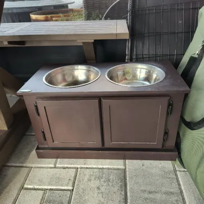 Elevated wooden dog feeder station in a dark brown finish, featuring two stainless steel bowls and a two-door storage cabinet below.