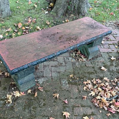 Weathered concrete garden bench featuring a red-painted rectangular top with a decorative scalloped border, supported by two...