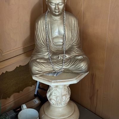 Gold-painted seated Buddha statue adorned with a silver bead necklace, displayed on a decorative pedestal stand.
