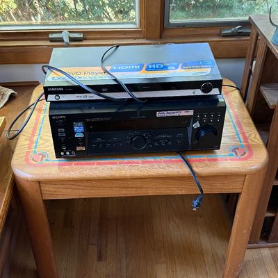 Samsung DVD player stacked on top of a Sony audio/video control center receiver, resting on a small wooden side table with a decorative...