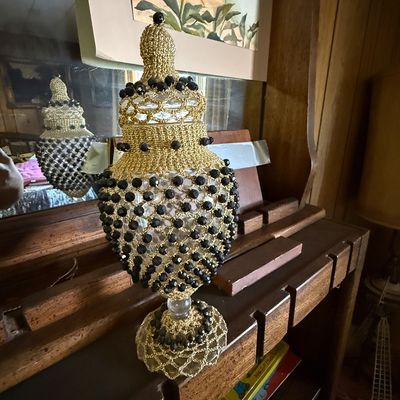 Vintage glass apothecary jar with an ornate, handmade gold crochet and black beaded cover, displayed on a wooden shelf.