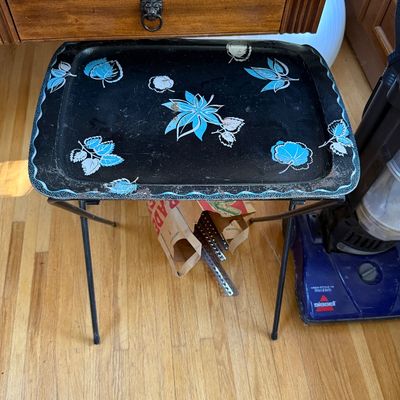 Vintage mid-century folding TV tray table featuring a black metal top adorned with blue and white floral and leaf motifs.