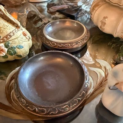Pair of vintage pewter or silver-plated coasters with ornate scrollwork borders, displayed alongside various ceramic and glass collectibles.