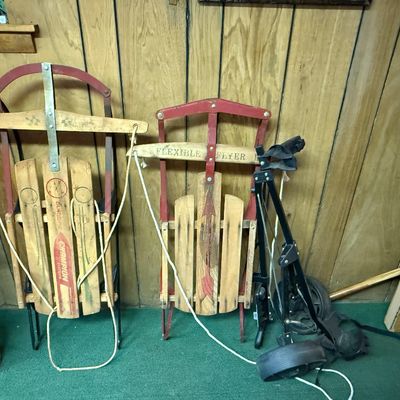Two vintage wooden snow sleds, including a Gladding Champion and a Flexible Flyer, alongside a black folding golf pull cart.