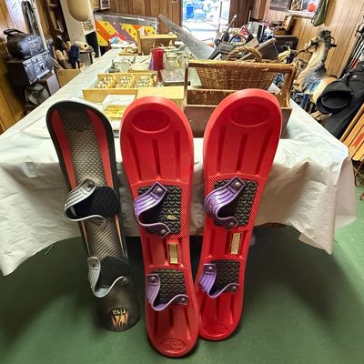Vintage plastic snowboards, including a black Riva model and a pair of red Sledz snowboards with purple bindings, leaning against a table.