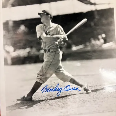 Vintage black and white photograph of baseball player Mickey Owen in mid-swing, featuring a blue ink signature across the bottom.