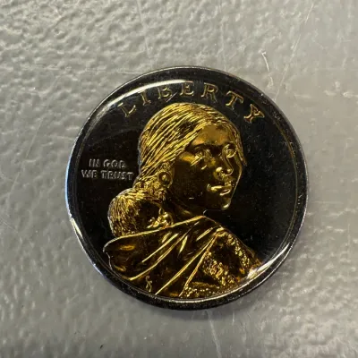 Sacagawea golden dollar coin featuring a gold-plated relief of Sacagawea and her infant son, Jean Baptiste, against a dark background.