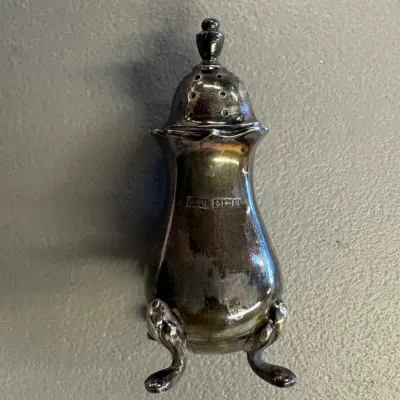 Vintage sterling silver pepper shaker featuring three cabriole legs, a domed perforated lid, and visible hallmarks on the body.