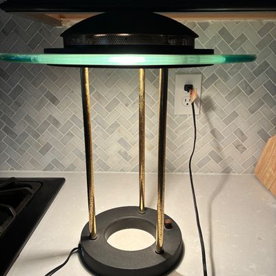 Sale Photo Thumbnail #552: Postmodern halogen desk lamp featuring a round black base, three brass-tone columnar supports, a clear thick glass disc diffuser collar, and a black dome top with perforated ventilation mesh. Powers on and lights up as shown. Shows light age-appropriate p