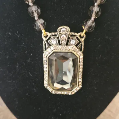Sale Photo Thumbnail #192: This piece is an authentic Heidi Daus Art Deco-inspired pendant necklace. It features a large, emerald-cut smoky glass crystal housed in an intricate, crystal-encrusted bronzetone setting, paired with a strand of faceted smoky gray glass accent beads