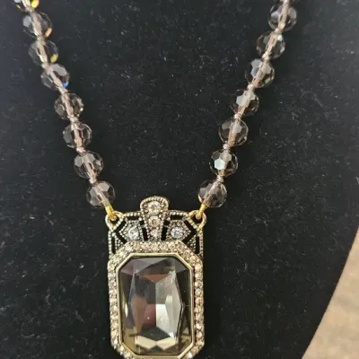 Sale Photo Thumbnail #191: This piece is an authentic Heidi Daus Art Deco-inspired pendant necklace. It features a large, emerald-cut smoky glass crystal housed in an intricate, crystal-encrusted bronzetone setting, paired with a strand of faceted smoky gray glass accent beads