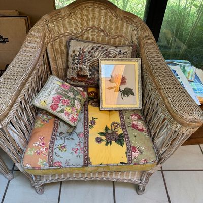 Vintage wicker armchair with a colorful floral patchwork cushion, styled with two decorative throw pillows and a framed needlepoint of a...