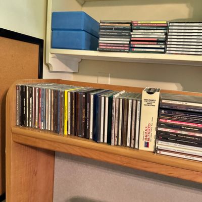 Large collection of classical and theatrical CDs in jewel cases, organized on a wooden shelf alongside two blue yoga blocks.