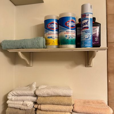Assorted household cleaning supplies including Clorox wipes and Lysol spray on a wooden wall shelf, with folded bath towels stacked on...