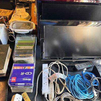 Assorted electronics and office supplies, including a Sharp flat-screen monitor, a power strip, various cables, and a stack of Memorex...