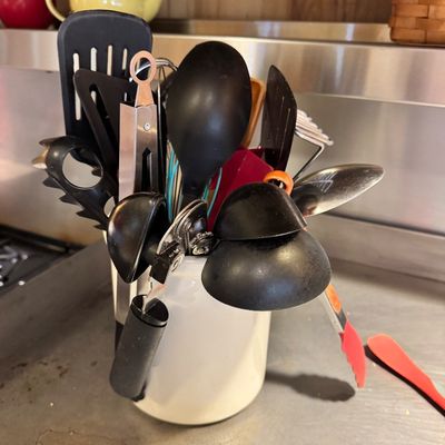 Assorted kitchen utensils, including spatulas, spoons, tongs, and a can opener, stored in a white ceramic crock.
