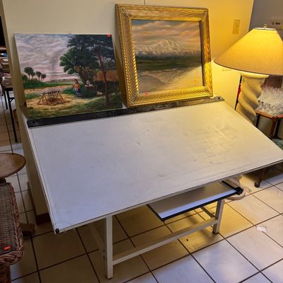 Vintage drafting table with a white top and metal base, displaying two framed landscape paintings.