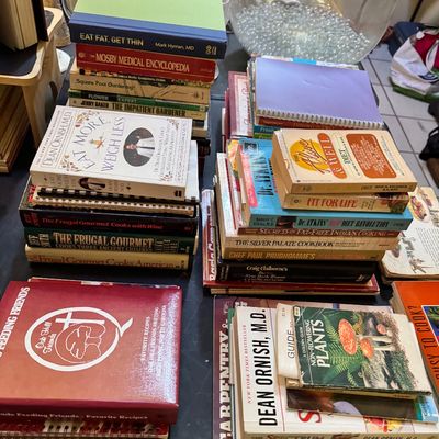 Assorted vintage and modern books, including cookbooks, gardening guides, and health and diet books, arranged in stacks on a table.