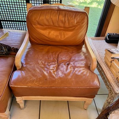 Vintage French provincial style armchair featuring a light-colored wood frame and worn tan leather cushions.