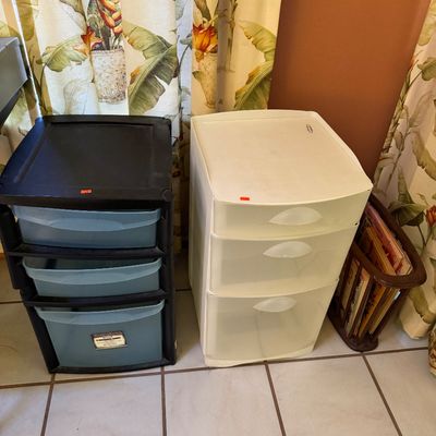 Two plastic three-drawer storage carts, one black with blue translucent drawers and one solid white, shown alongside a small wooden...